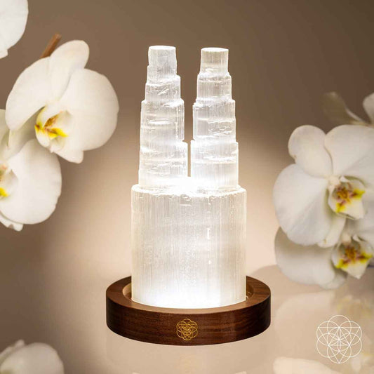 Two-tower selenite lamp with two lit crystal columns on wooden base near orchids