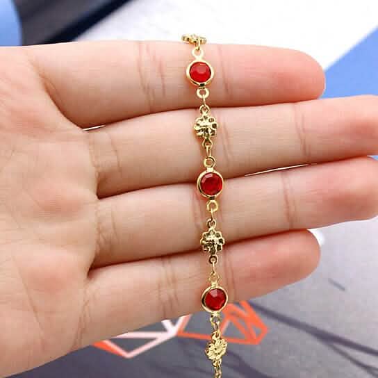 Gold-tone anklet bracelet held in hand, red crystal bezel links and flower charms