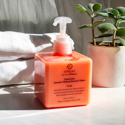 Orange facial cleanser pump bottle with white pump, on countertop beside towel and plant pot.