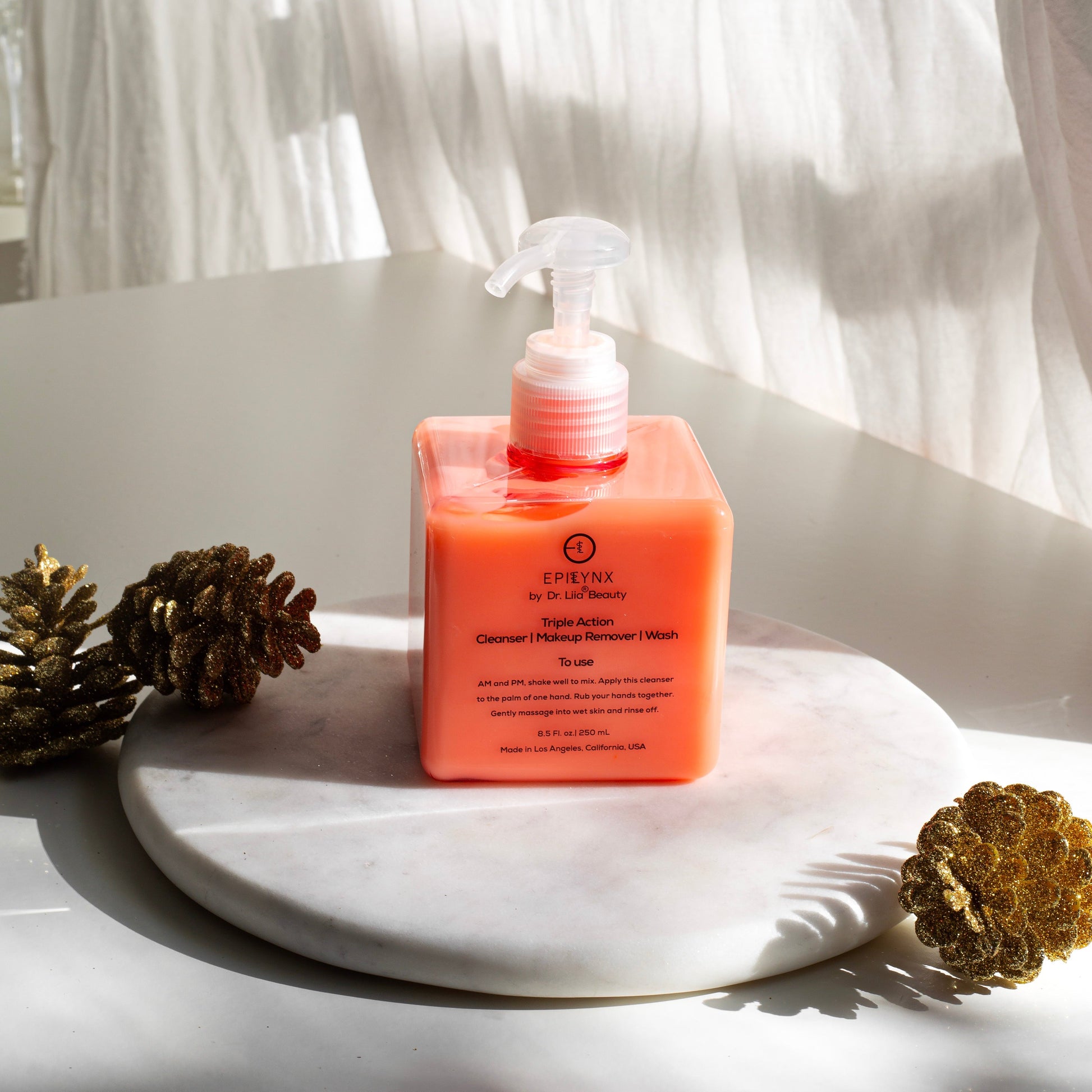 Orange facial cleanser pump bottle centered on white pedestal with decorative pinecones.