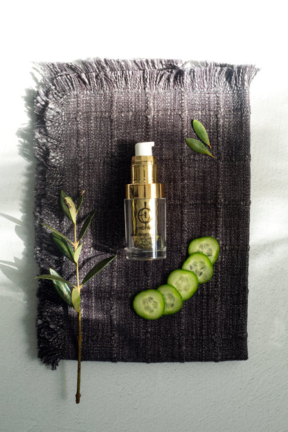 Serum bottle with gold pump on a dark towel with cucumber slices.