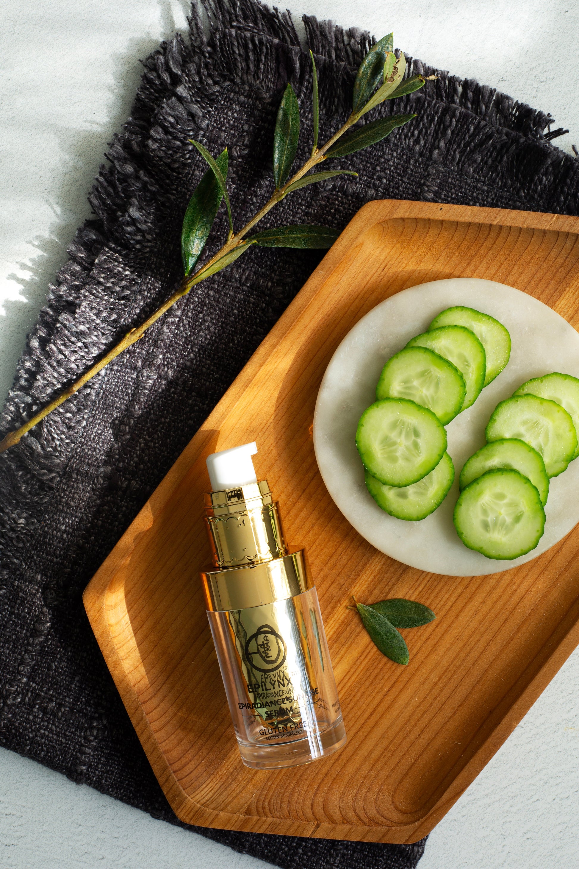 Serum bottle with gold pump on a wooden tray beside cucumber slices.
