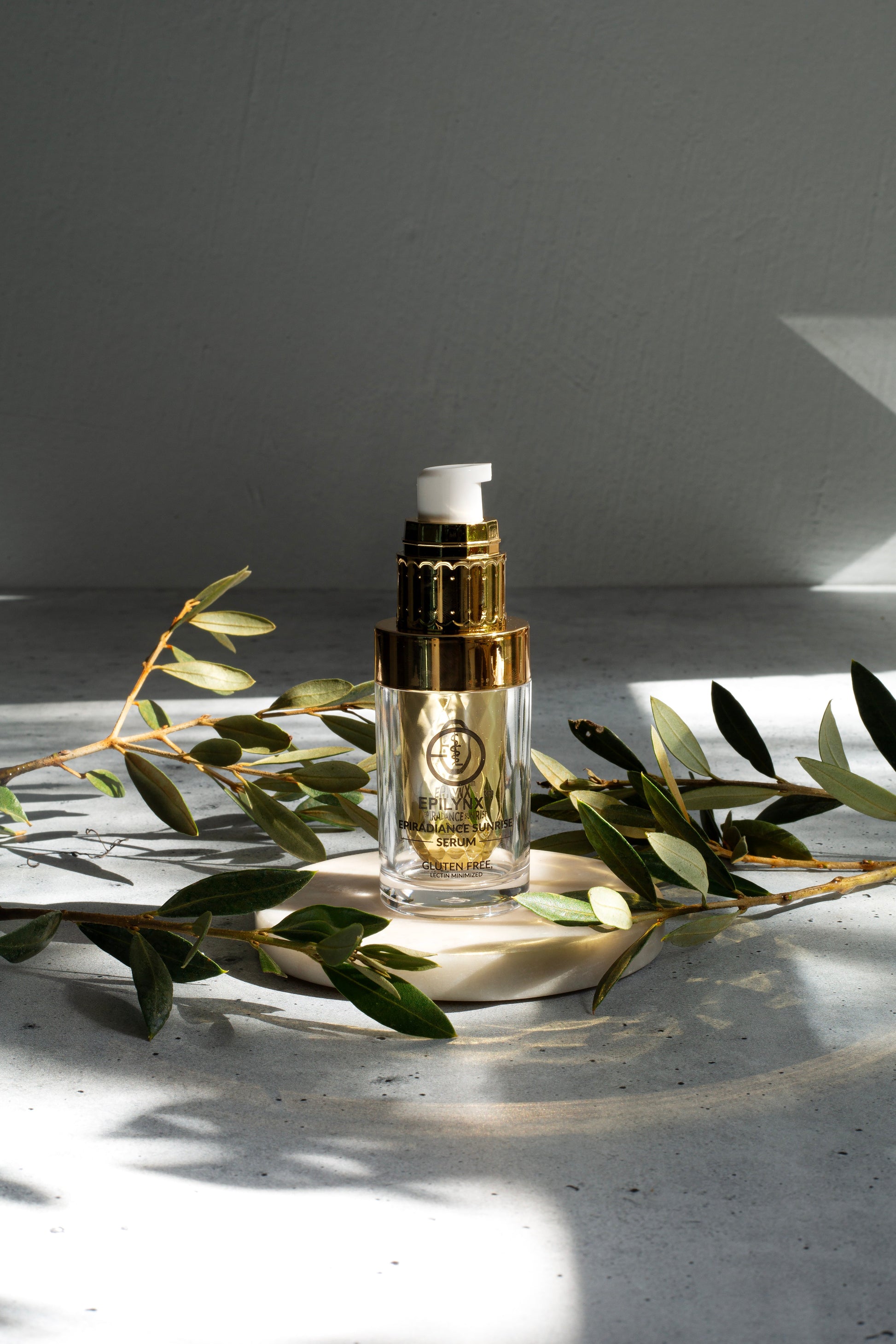Glass serum bottle with gold pump on a wooden base with green leaf branches.