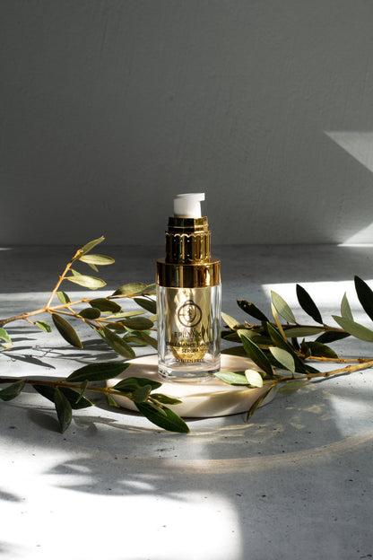 Glass serum bottle with gold pump on a wooden base with green leaf branches.
