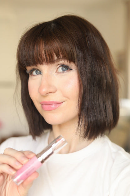 Pink lip gloss wand in clear tube, held near lips with natural rosy shine