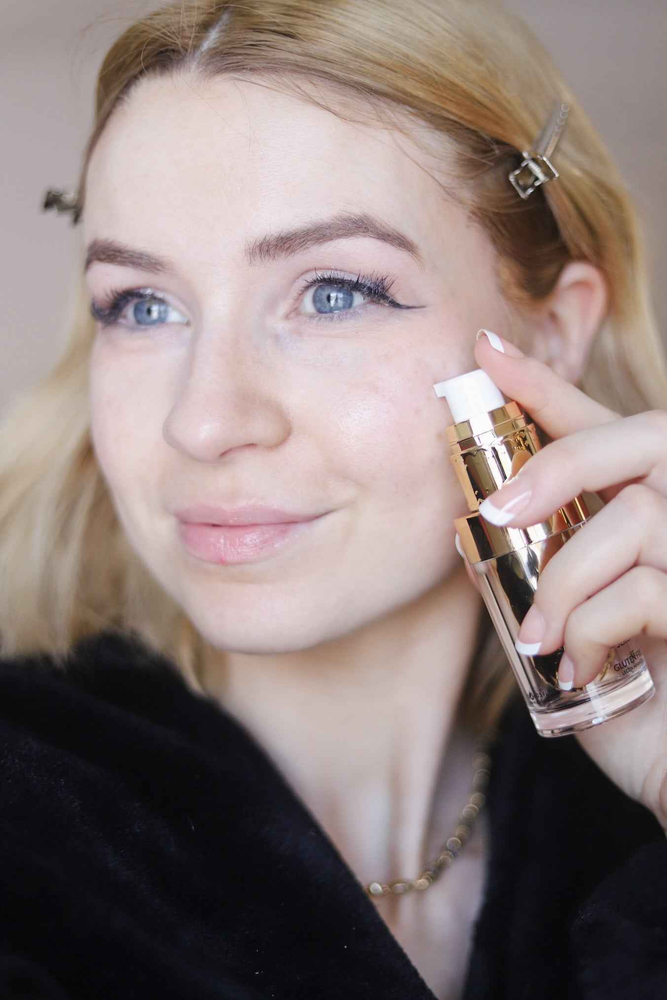 Woman holding a serum bottle while applying pale gel near her cheek.