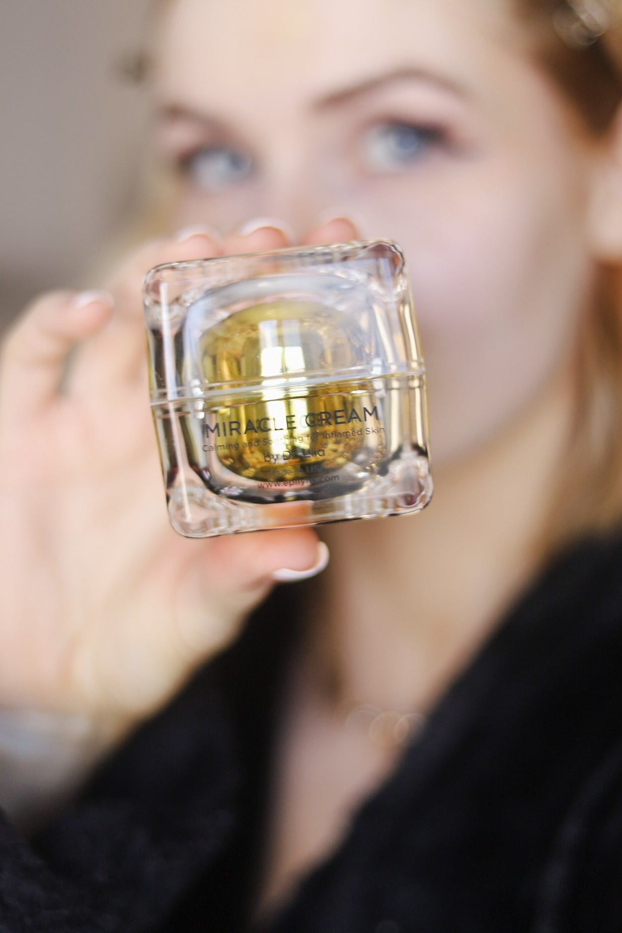 Person holding a clear Miracle Cream jar with gold-yellow gel near cheek