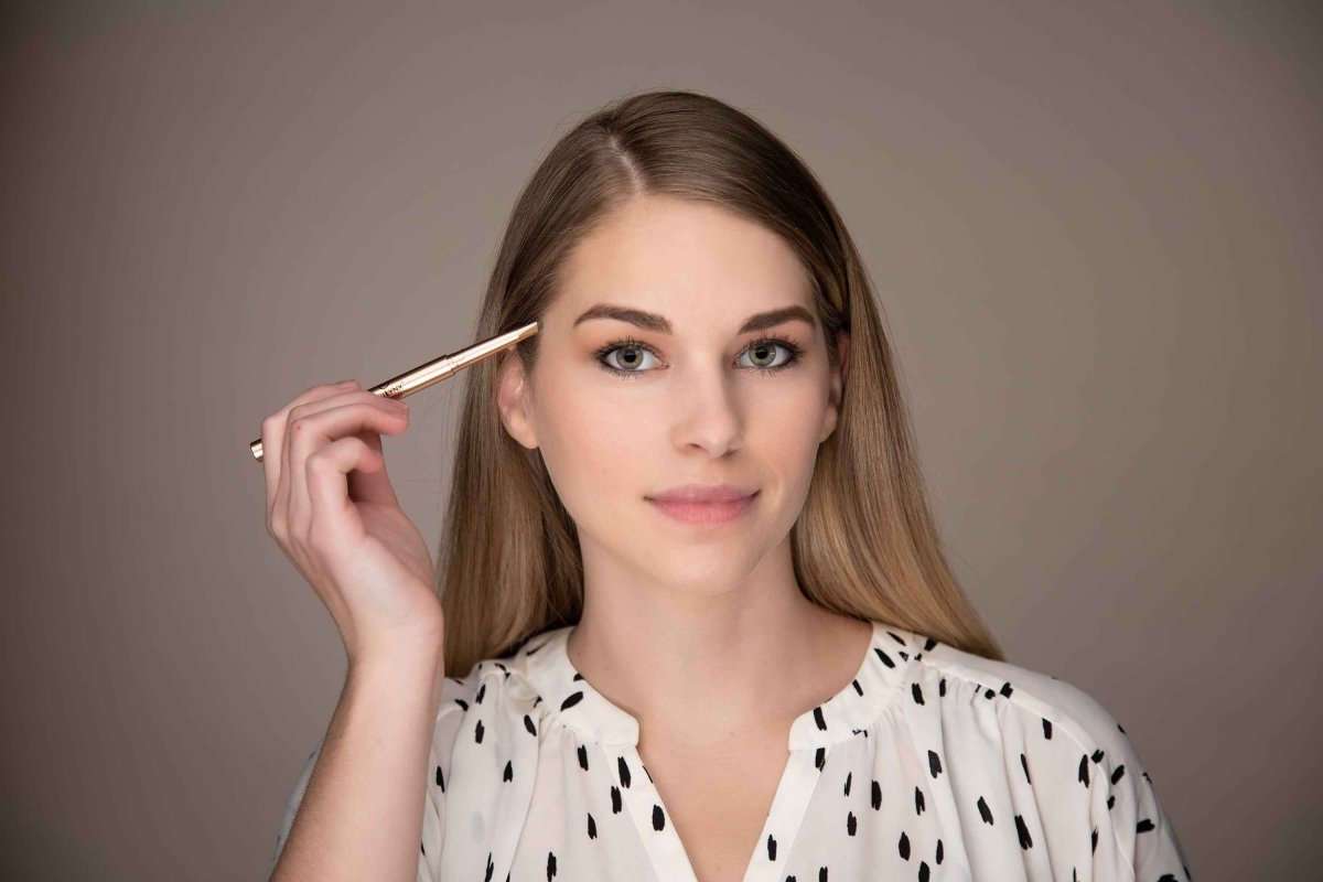 Woman holding an eyebrow pencil above her face; tip poised for brow definition.