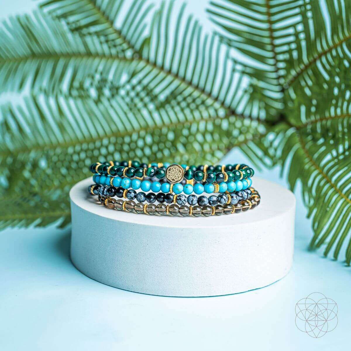 Four beaded bracelets on a light pedestal, turquoise and dark beads with a round gold center charm