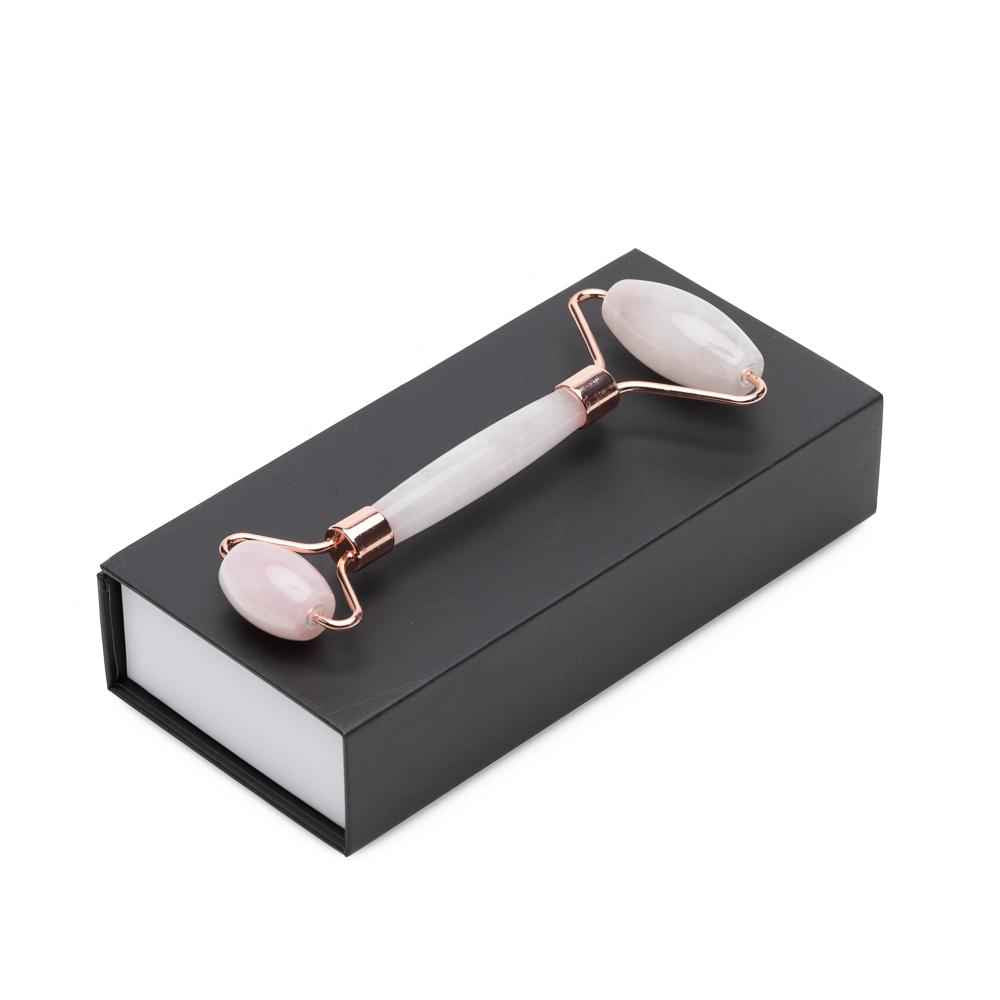 Dual-ended facial roller with pink quartz heads and rose-gold frames on black box