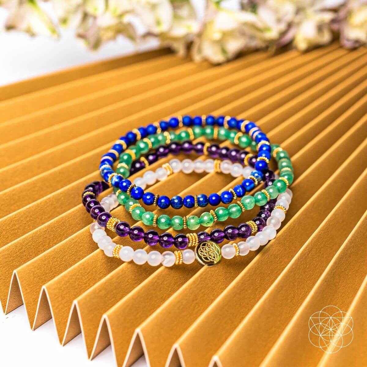 Four beaded bracelets with gold charm on wooden tray, blue, green, purple and white beads