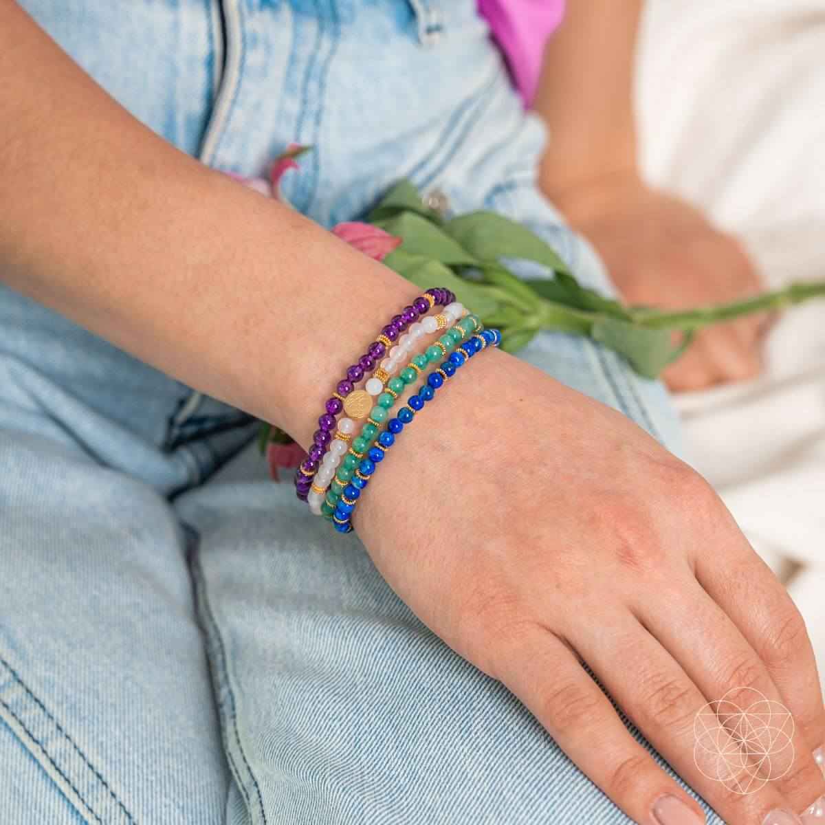 Stack of four beaded bracelets with gold charm on wrist, purple, blue, green and white beads
