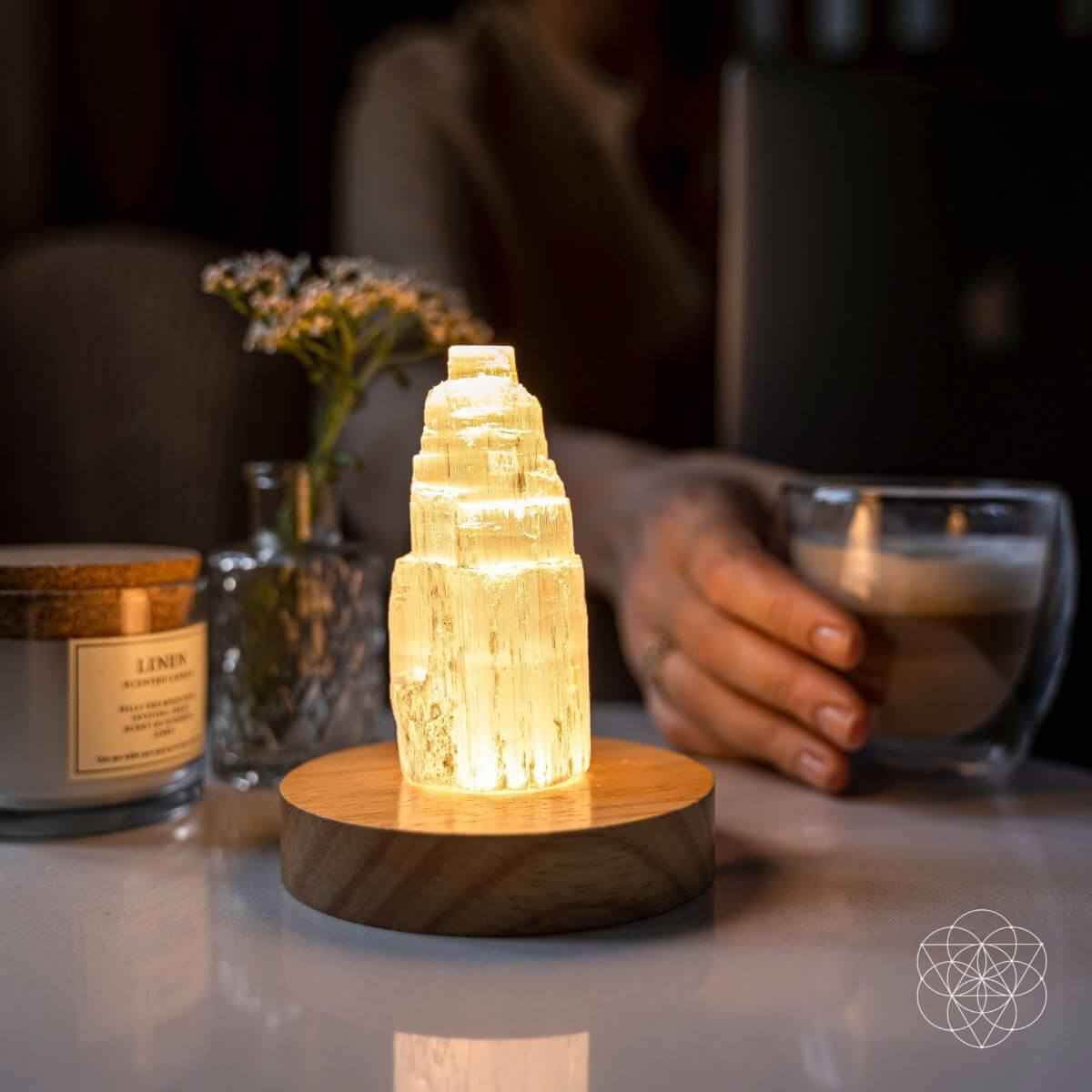 Selenite cleansing lamp glowing amber crystal on round wooden base, hand beside