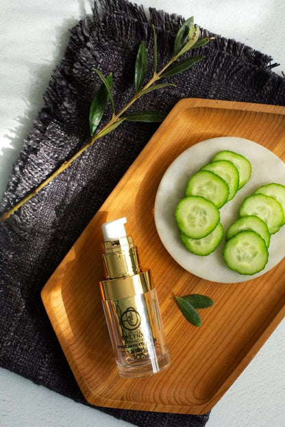 Facial serum bottle with gold pump on wooden tray with cucumber slices and herbs.