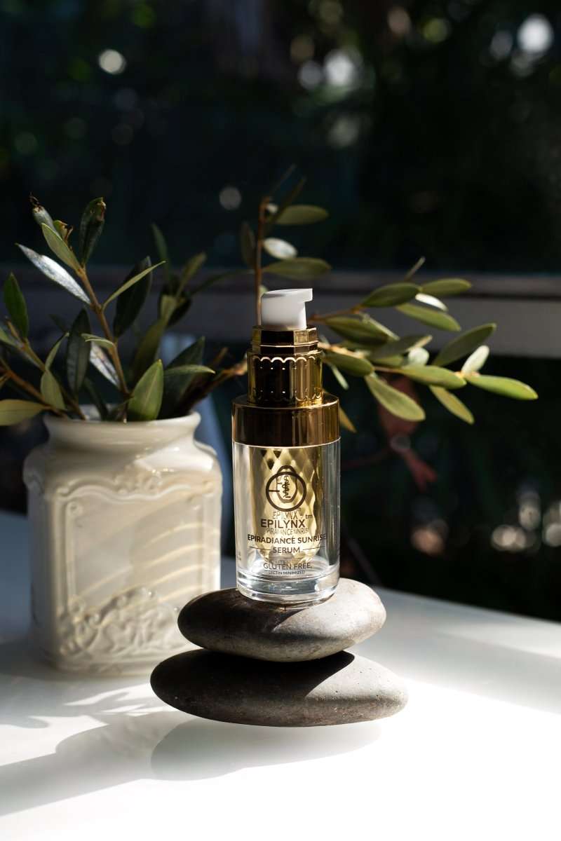 Gold-pump serum bottle on dark stone with greenery beside it.