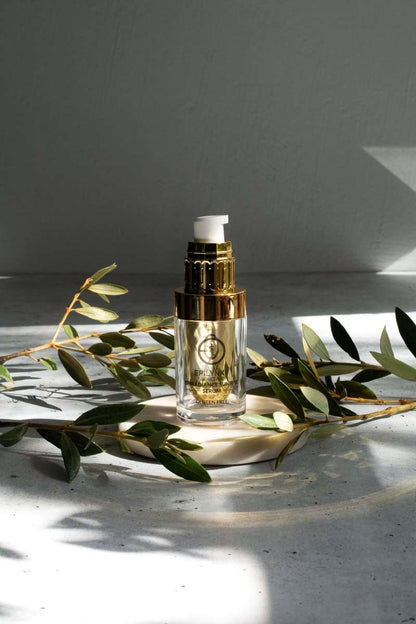 Facial serum bottle with gold pump on stone surface with olive leaves.