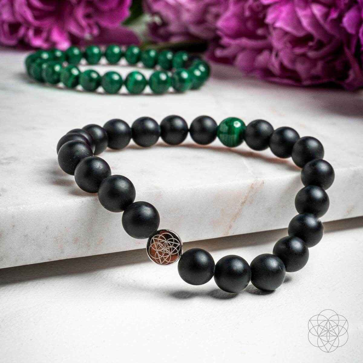 Two beaded bracelets laid flat: matte black beads and green accents with geometric charm