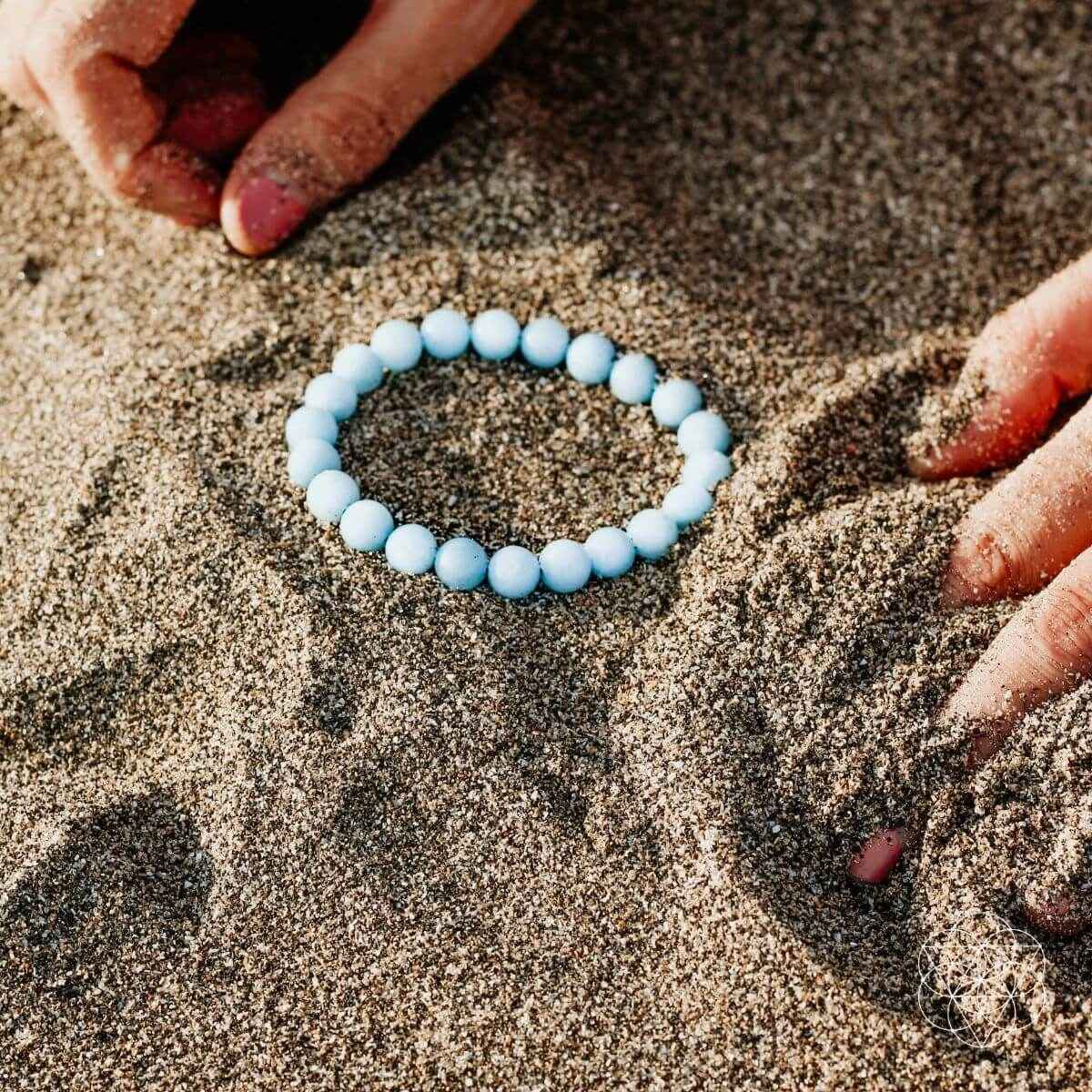 Sky-blue beaded bracelet laid on sand, round bead strand.