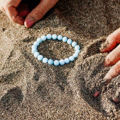 Sky-blue beaded bracelet laid on sand, round bead strand.