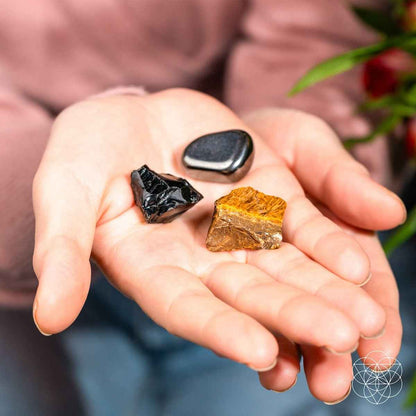 Hand holding protection stones: black shard, dark gray smooth stone, and gold-brown piece.
